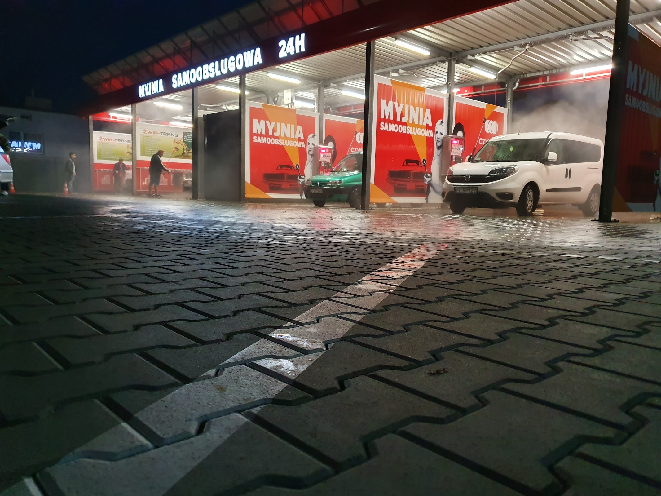 Myjnia Jedynka nocą - podświetlone stanowiska, dwa auta w trakcie mycia, mokra kostka brukowa.